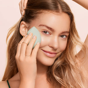 Teal teardrop makeup blending sponge held to apply foundation on cheek