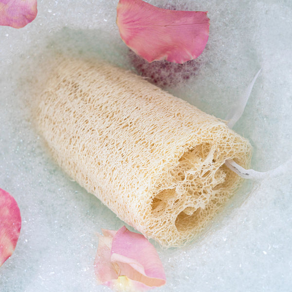 Loofah body sponge rolled up for use, with bath bubbles and petals nearby