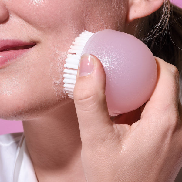 Pink facial cleansing brush held against cheek for scrubbing
